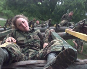 Exhausted female soldier sprawls on horizontal ladder obstacle while other soldiers climb over it in background.